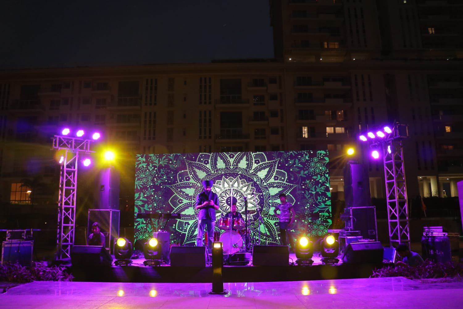 Live band performing outdoors under string lights with green mandala backdrop